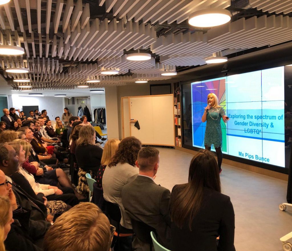 Pips Bunce standing on stage hosting a session in front of a full auditorium, the slide she is talking to is on the topic of exploring the spectrum of Gender Diversity and LGBTQI+ identities