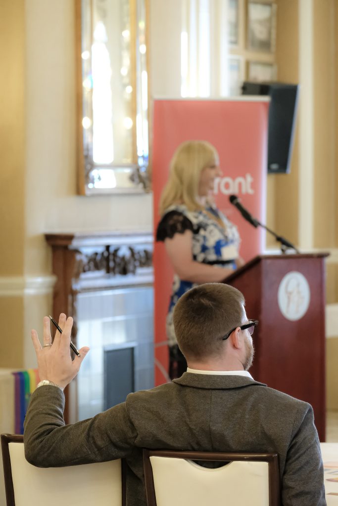 Pips Bunce standing behind a podium as part of a diversity conference, with members of the audience raising their hands with questions to ask